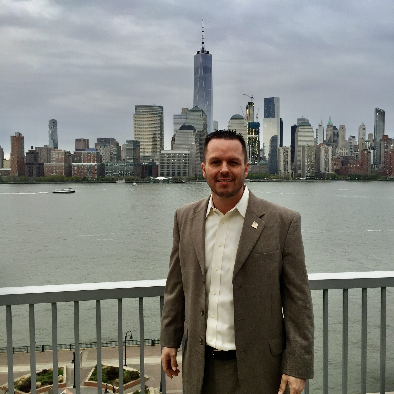 Jody Fuller in a tan sport coat on the Hyatt Regency Jersey City terrace with the Hudson River, lower Manhattan and One World Trade Center behind him
