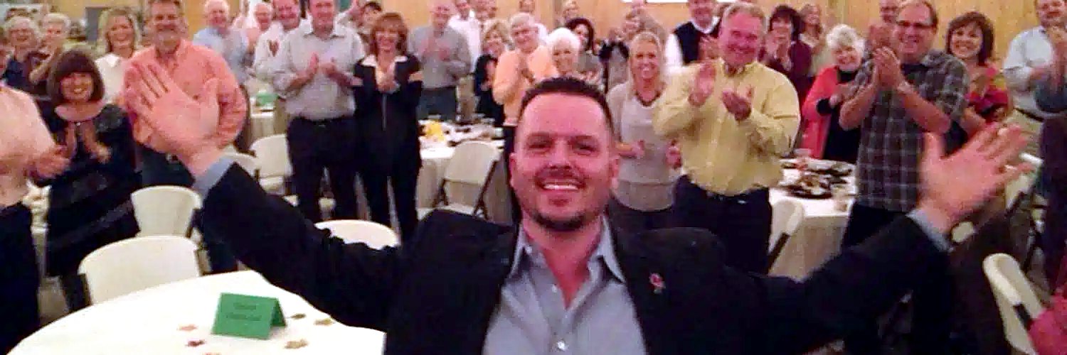 Jody Fuller with arms raised in front of a banquet audience giving him a standing ovation