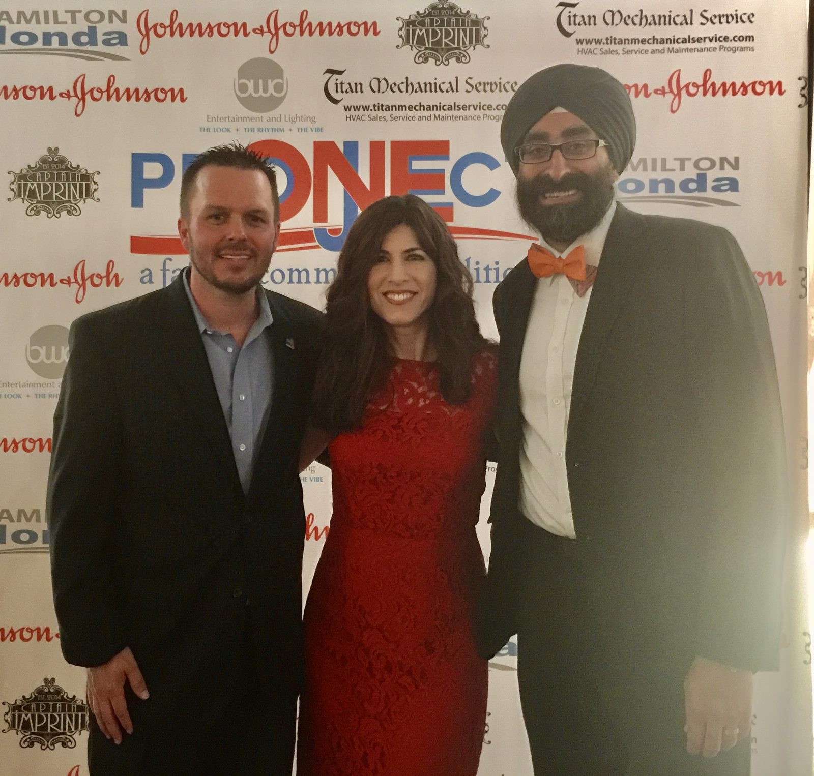 Jody Fuller with Amman Seehra in a turban and orange bow tie and a guest in a red dress in front of the PRONECT faith and community coalition sponsor backdrop